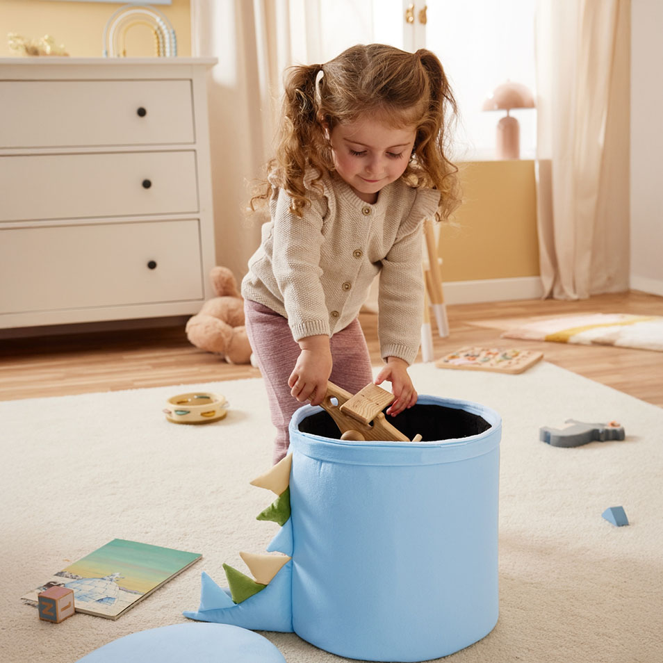 Dítě vkládá dřevěné hračky do modrého úložného koše s motivem dinosaura v ložnici.