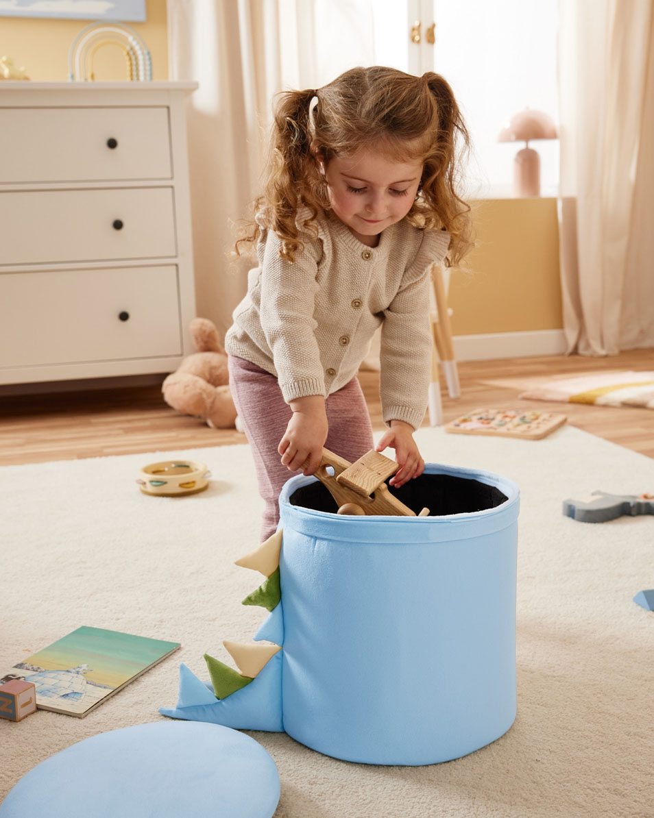 Dítě vkládá dřevěné hračky do modrého úložného koše s motivem dinosaura.