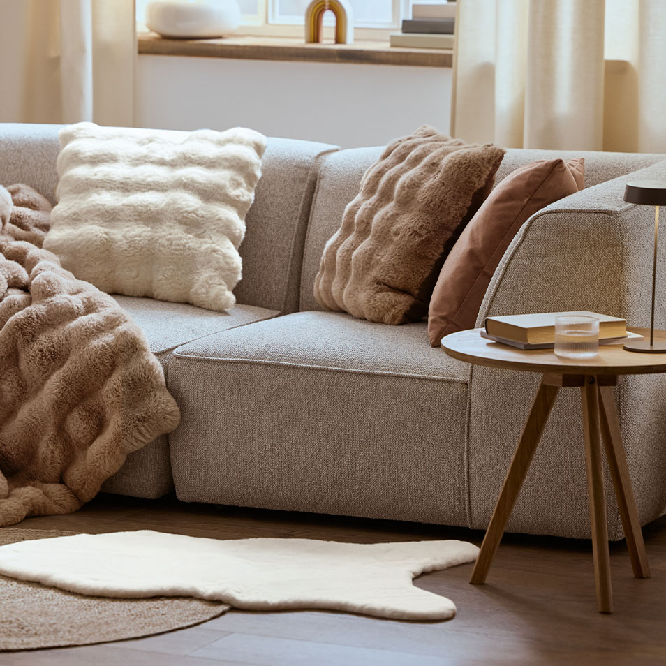 Šedá modulární pohovka s nadýchanými polštáři a přehozy, vedle konferenčního stolku.