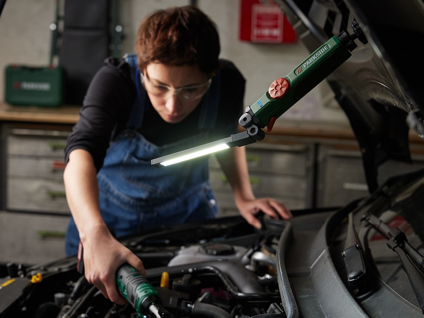 Mechanička používá přenosnou pracovní lampu Parkside k opravě motoru automobilu.