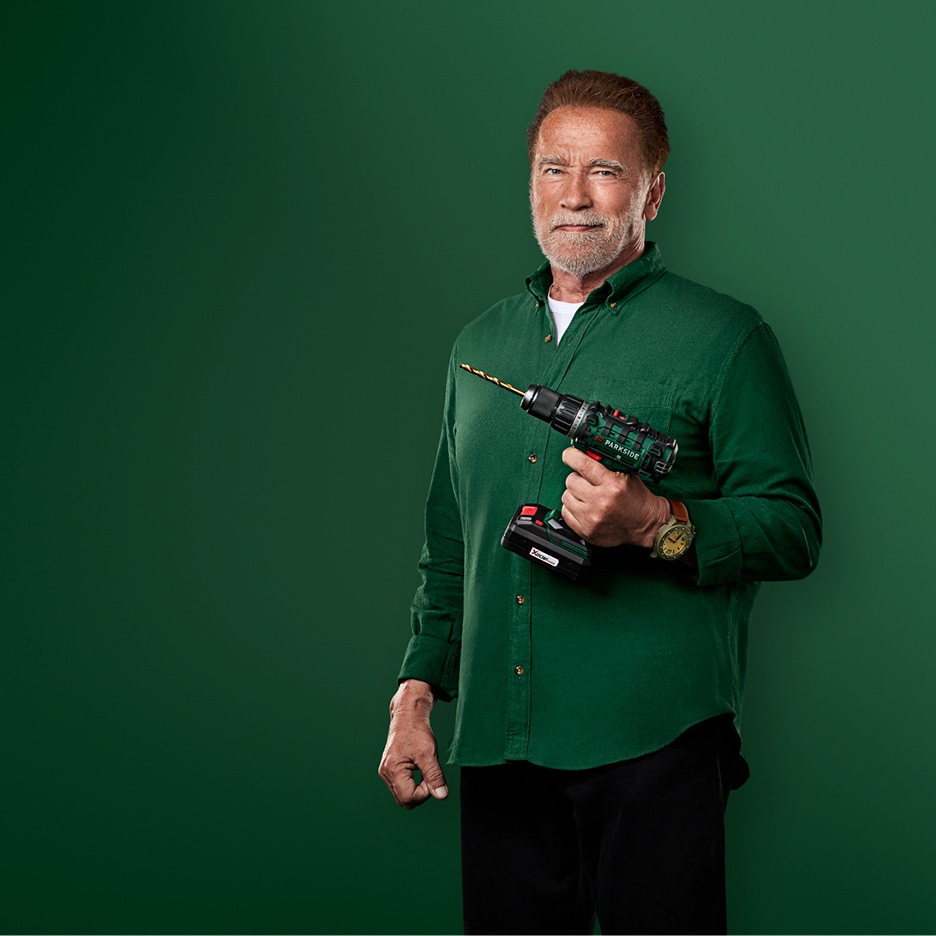 Arnold Schwarzenegger drží akumulátorovou vrtačku s vrtákem, oblečený v zelené košili.
