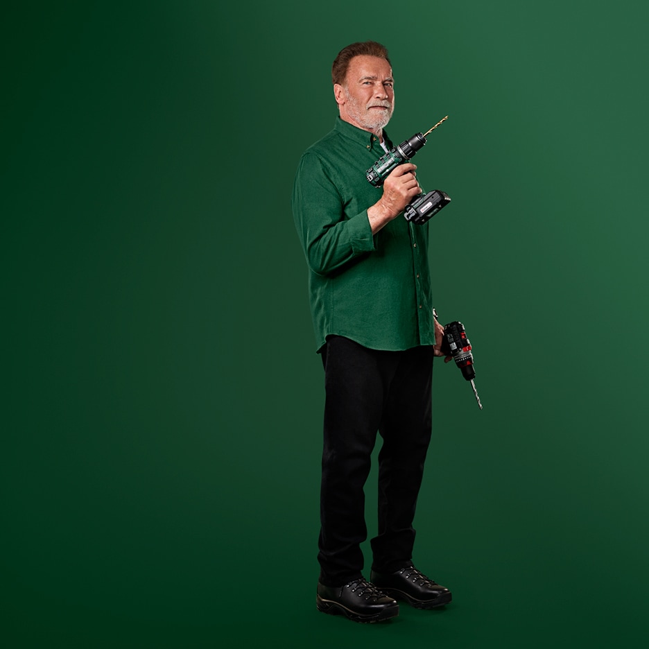 Arnold Schwarzenegger drží dva akumulátorové vrtací šroubováky na zeleném pozadí.