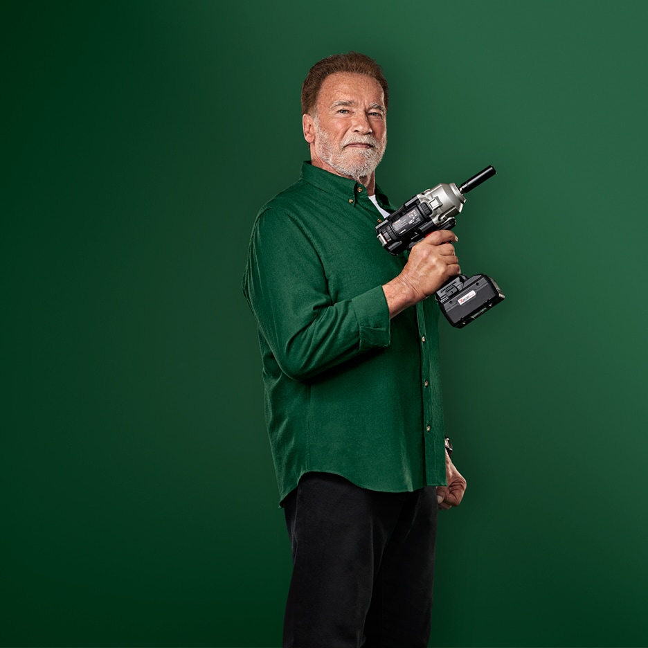 Arnold Schwarzenegger v zelené košili drží akumulátorový rázový utahovák.
