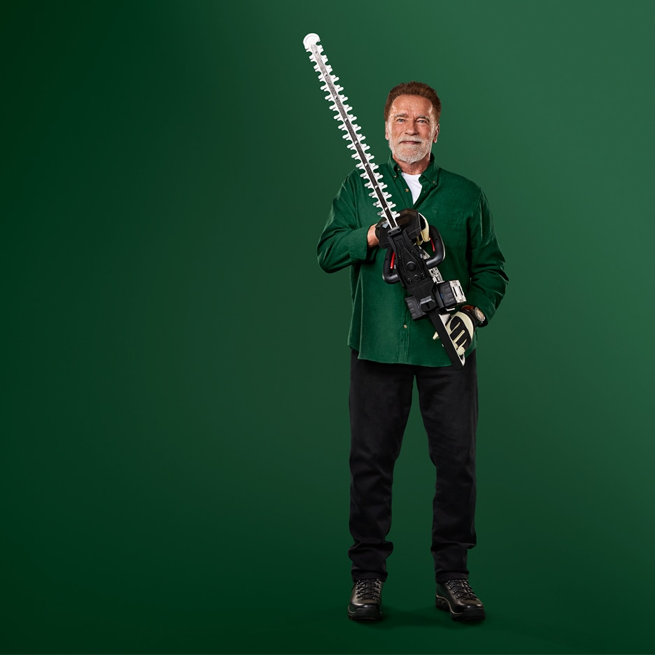 Arnold Schwarzenegger drží nůžky na živý plot, na sobě má zelenou košili a černé kalhoty.