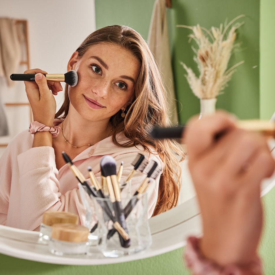 Žena si nanáší make-up štětcem před zrcadlem, s dalšími štětci a kosmetikou na poličce.