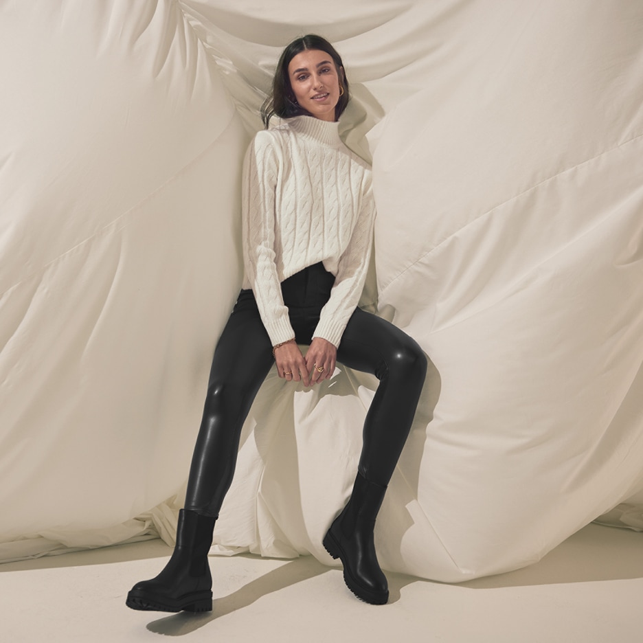 Žena v bílém pleteném svetru, černých legínách a černých botách sedí na bílé látce.