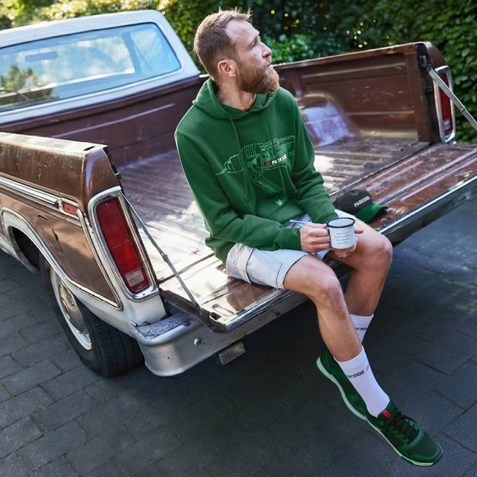 Muž v zelené mikině Parkside a šortkách sedí na korbě pickupu a drží hrnek.