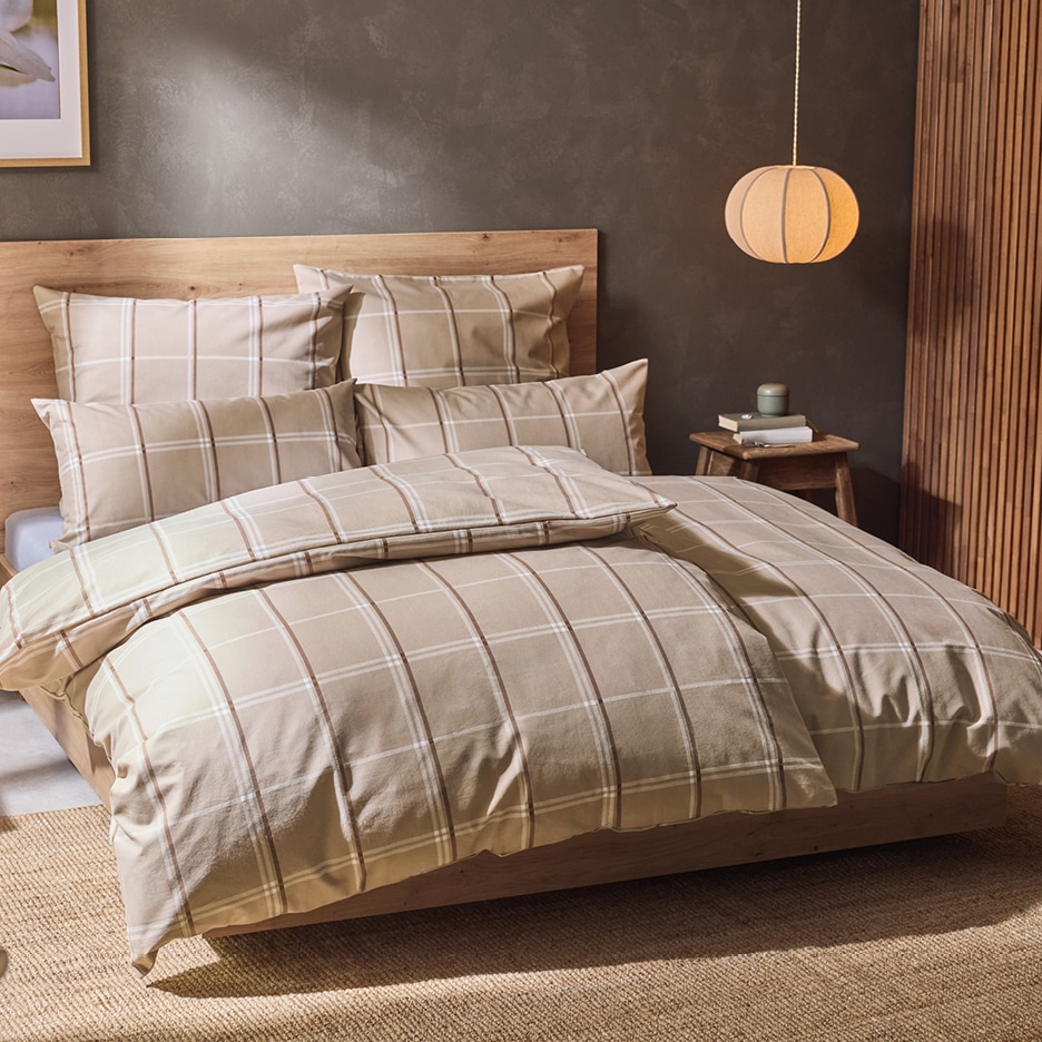 Béžové kostkované povlečení na dřevěné posteli v ložnici s lampou a nočním stolkem.