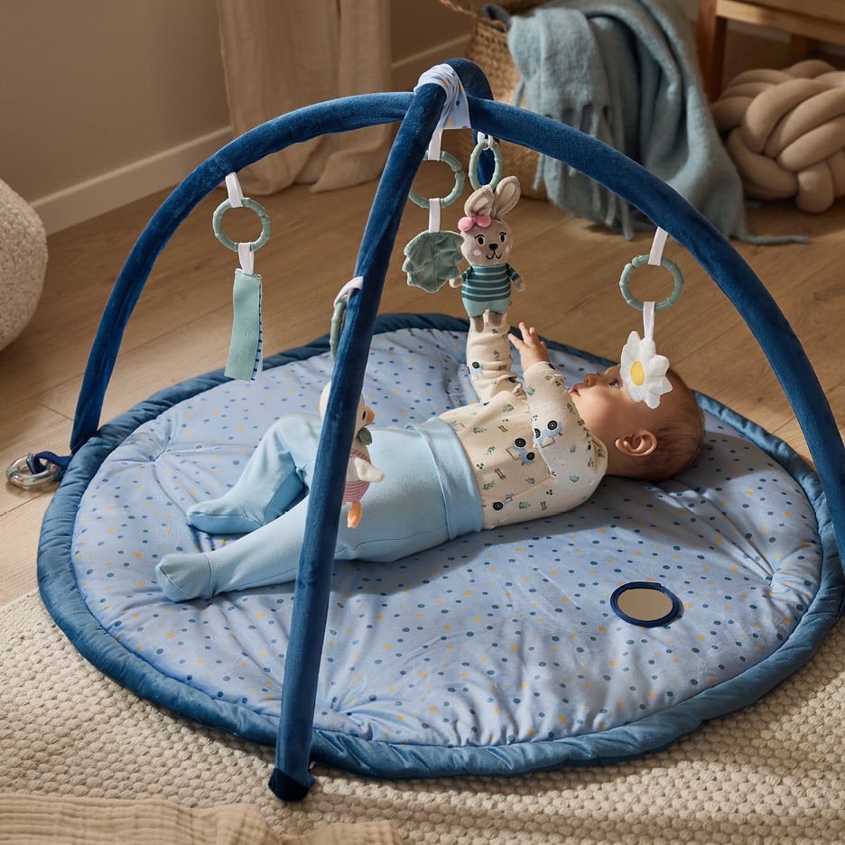 Dítě ležící na modré hrací podložce s hrazdičkou a zavěšenými hračkami.