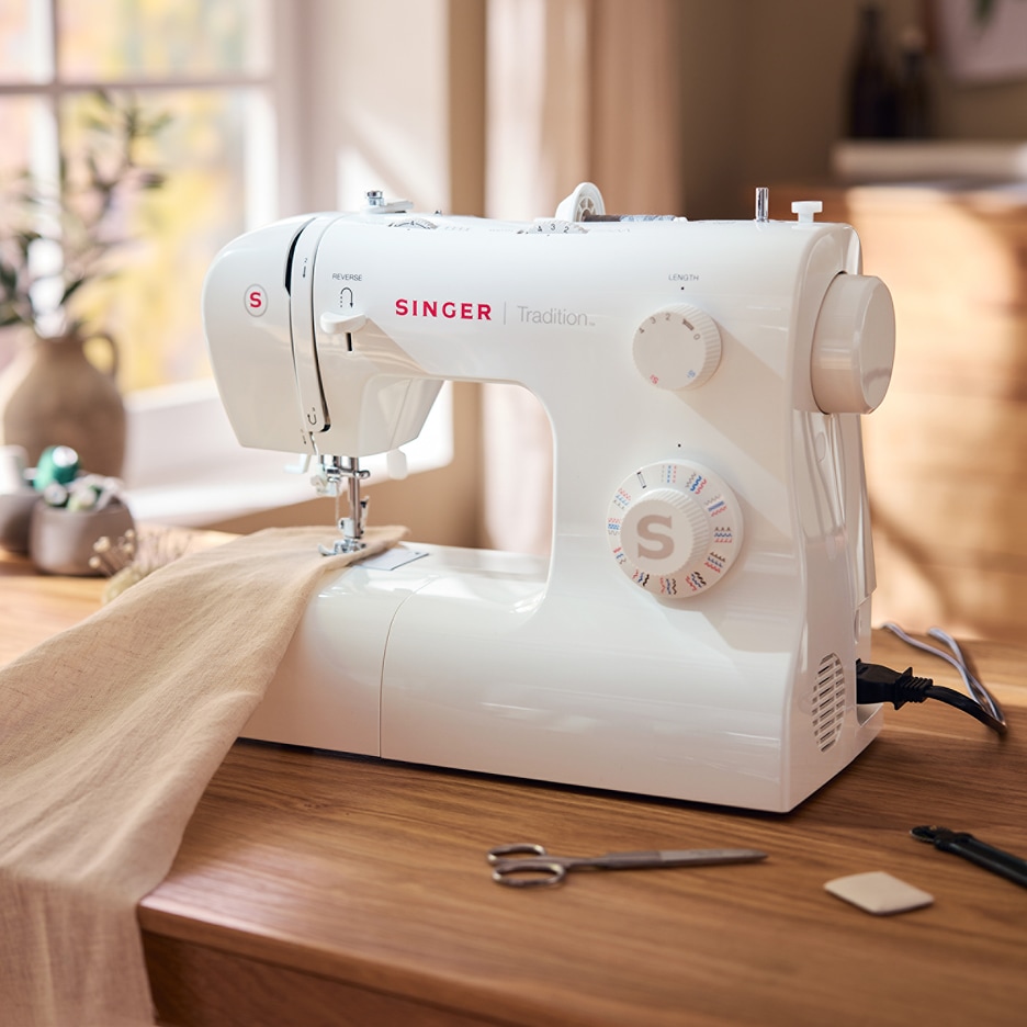 Bílý šicí stroj Singer Tradition na dřevěném stole s látkou a nůžkami.