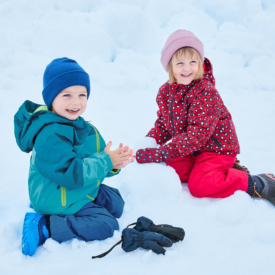 Dvě děti v zimních bundách a kalhotách si hrají ve sněhu, staví sněhuláka.
