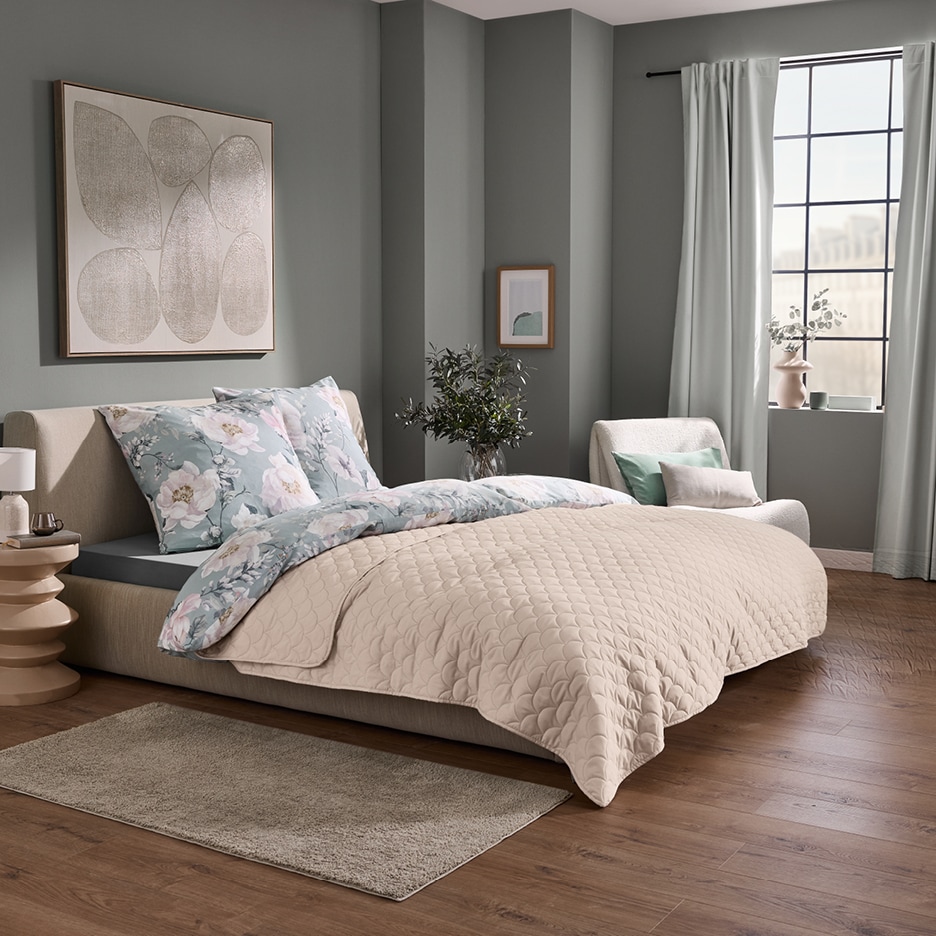 Ložnice s béžovou postelí, květinovým povlečením, béžovým přehozem a šedozelenými stěnami.