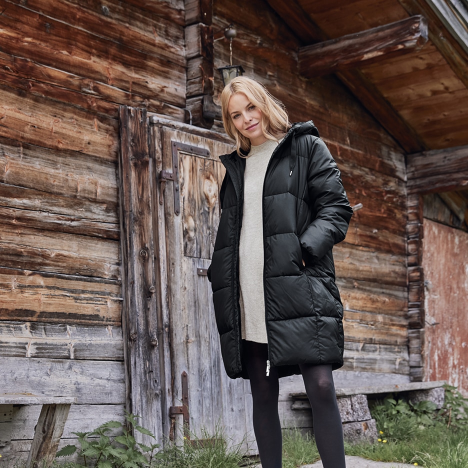 Žena v černé prošívané bundě a světlých šatech před dřevěnou chatou.