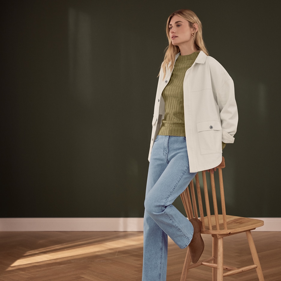 Žena v béžové bundě, zeleném svetru a džínách, opřená o dřevěnou židli.