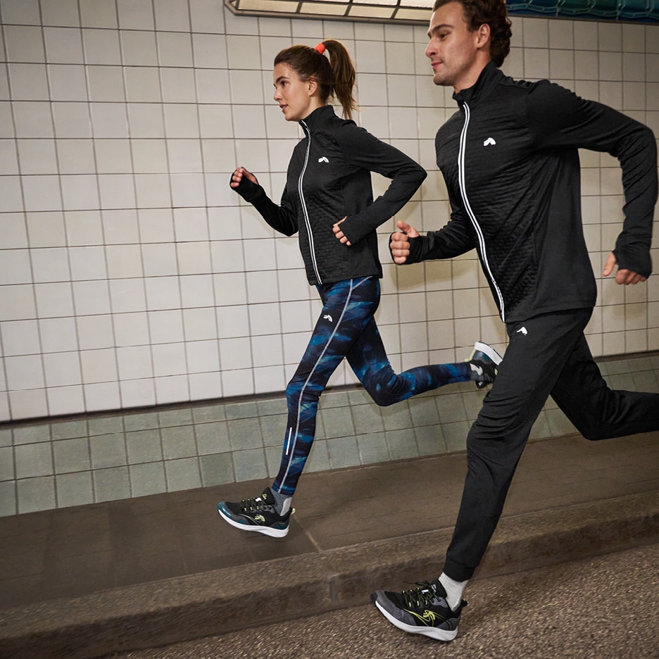 Dva běžci v černých sportovních bundách a kalhotách, žena v modrých legínách, běží v podchodu.