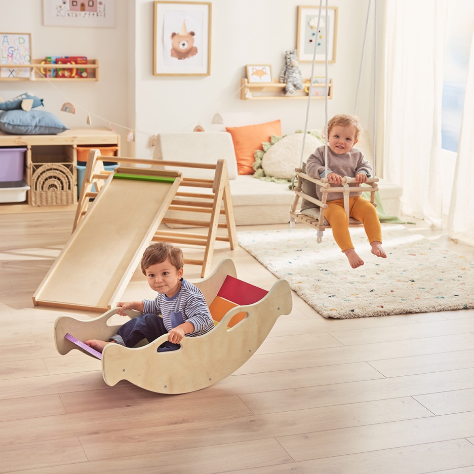 Dvě batolata si hrají v dětském pokoji s dřevěnou skluzavkou, houpačkou a houpací lodí.