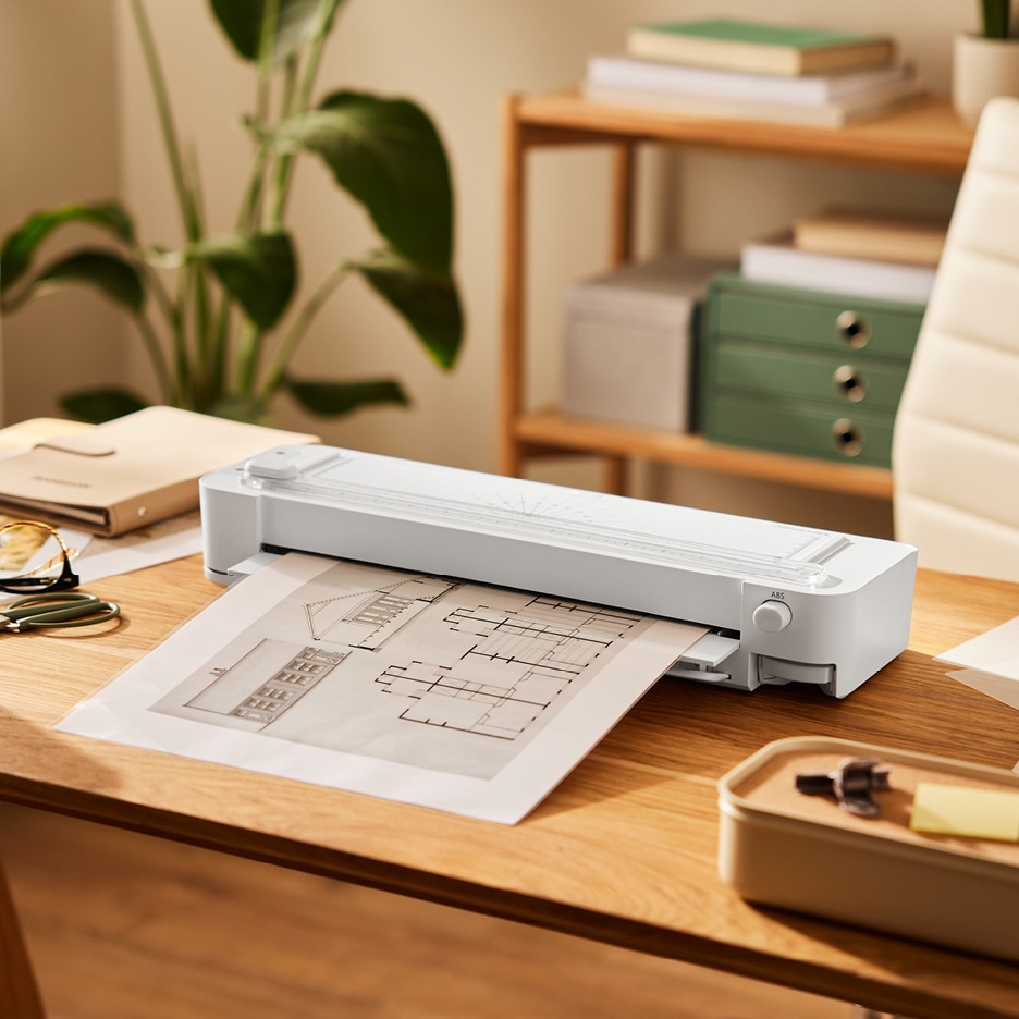 Laminátor na dřevěném stole s architektonickými plány, kancelářské potřeby a rostlina v pozadí.