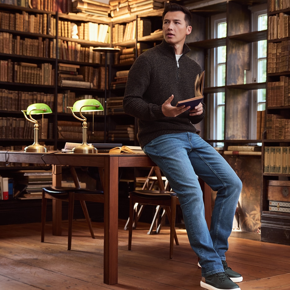 Muž v tmavě hnědém svetru a džínách sedí na stole v knihovně a čte knihu.