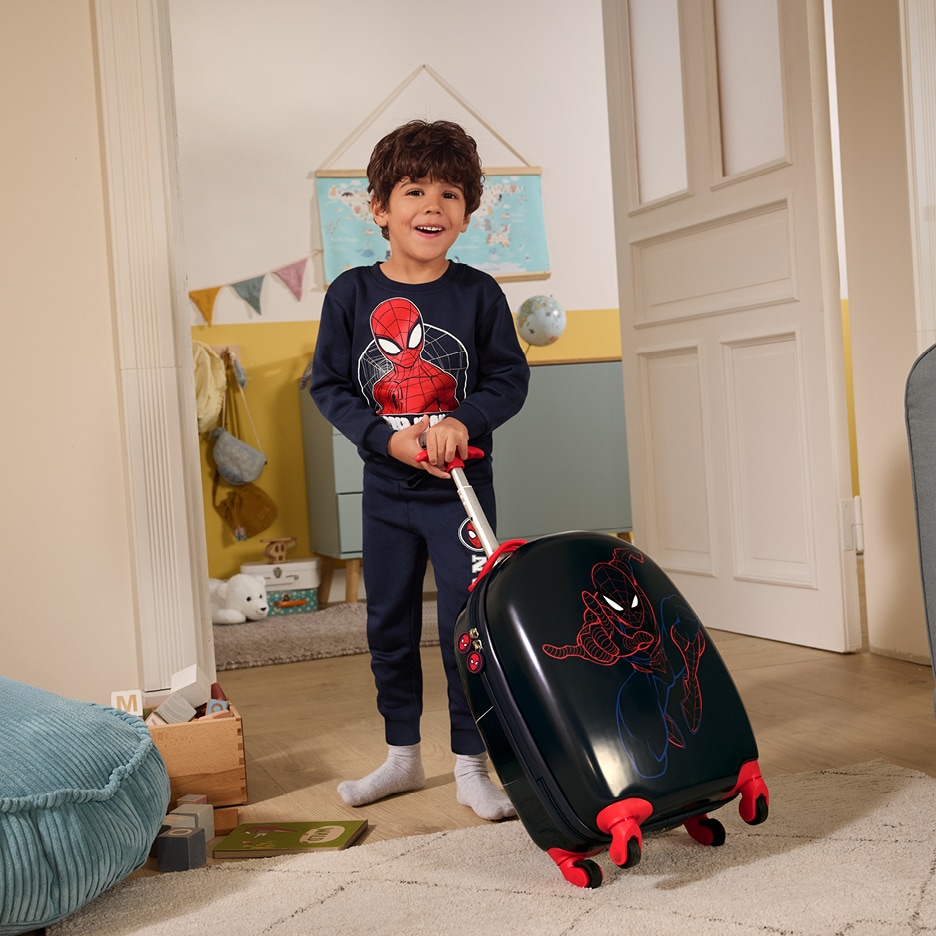 Chlapec v pyžamu s motivem superhrdiny táhne dětský kufr s motivem superhrdiny.