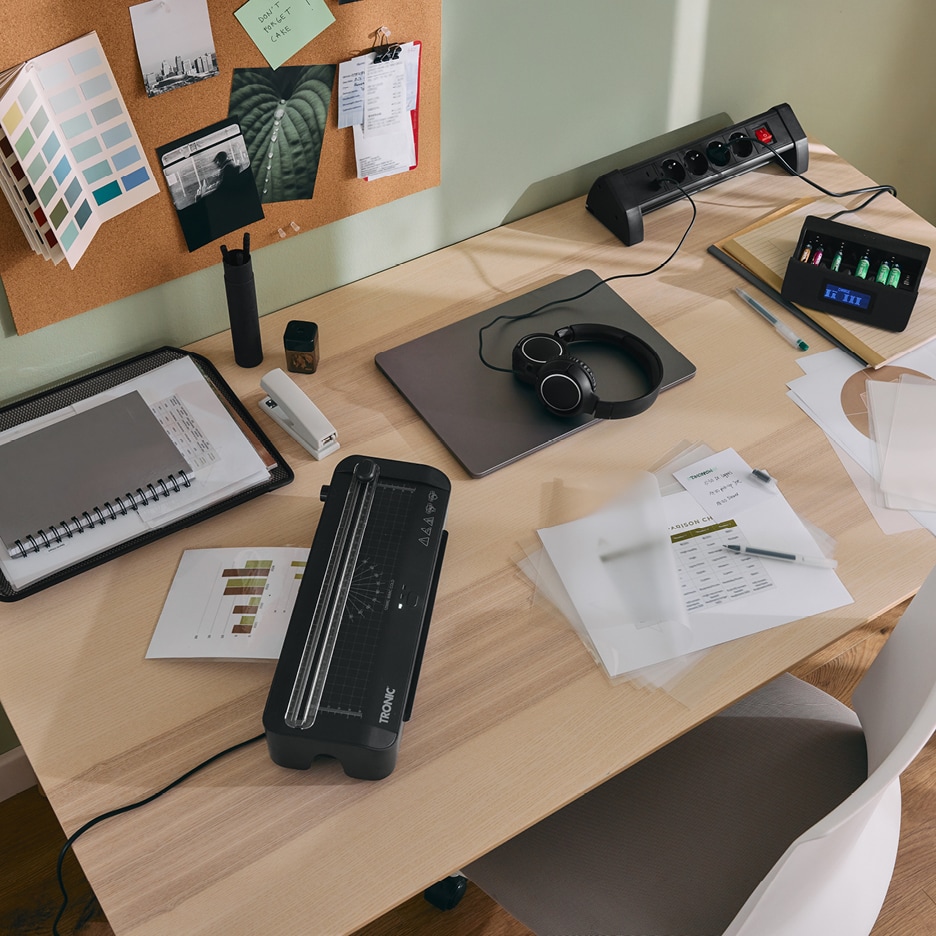 Pracovní stůl s laminátorem, notebookem, sluchátky, prodlužovacím kabelem a nabíječkou baterií.