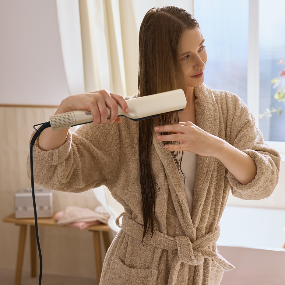 Žena v županu si žehlí mokré vlasy žehličkou na vlasy.