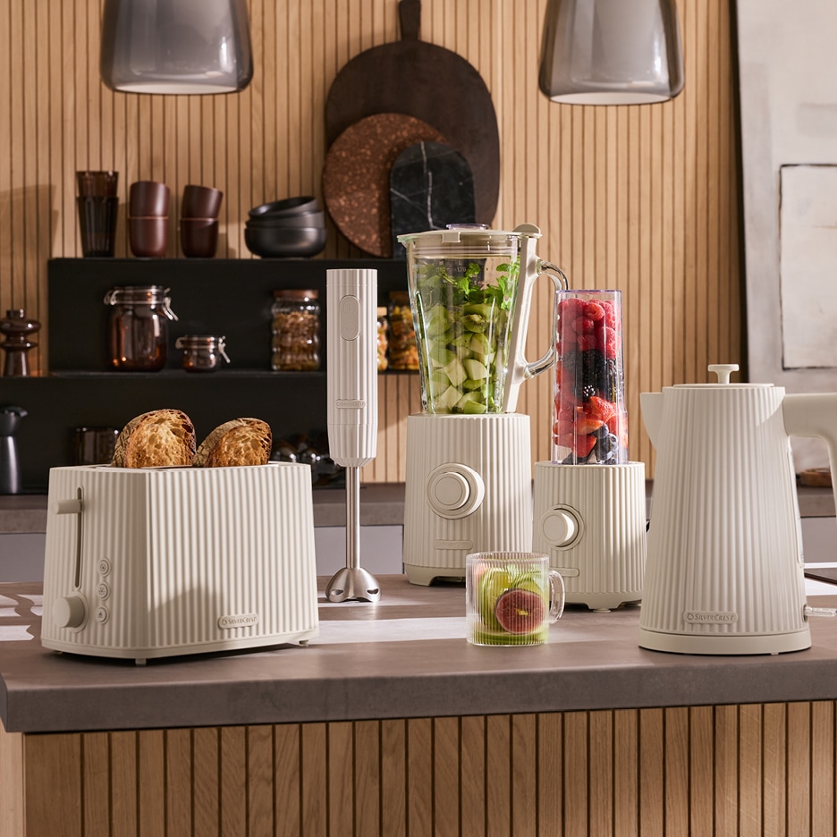 Kuchyňské spotřebiče v krémové barvě: toustovač, tyčový mixér, stolní mixér a rychlovarná konvice na kuchyňské lince.