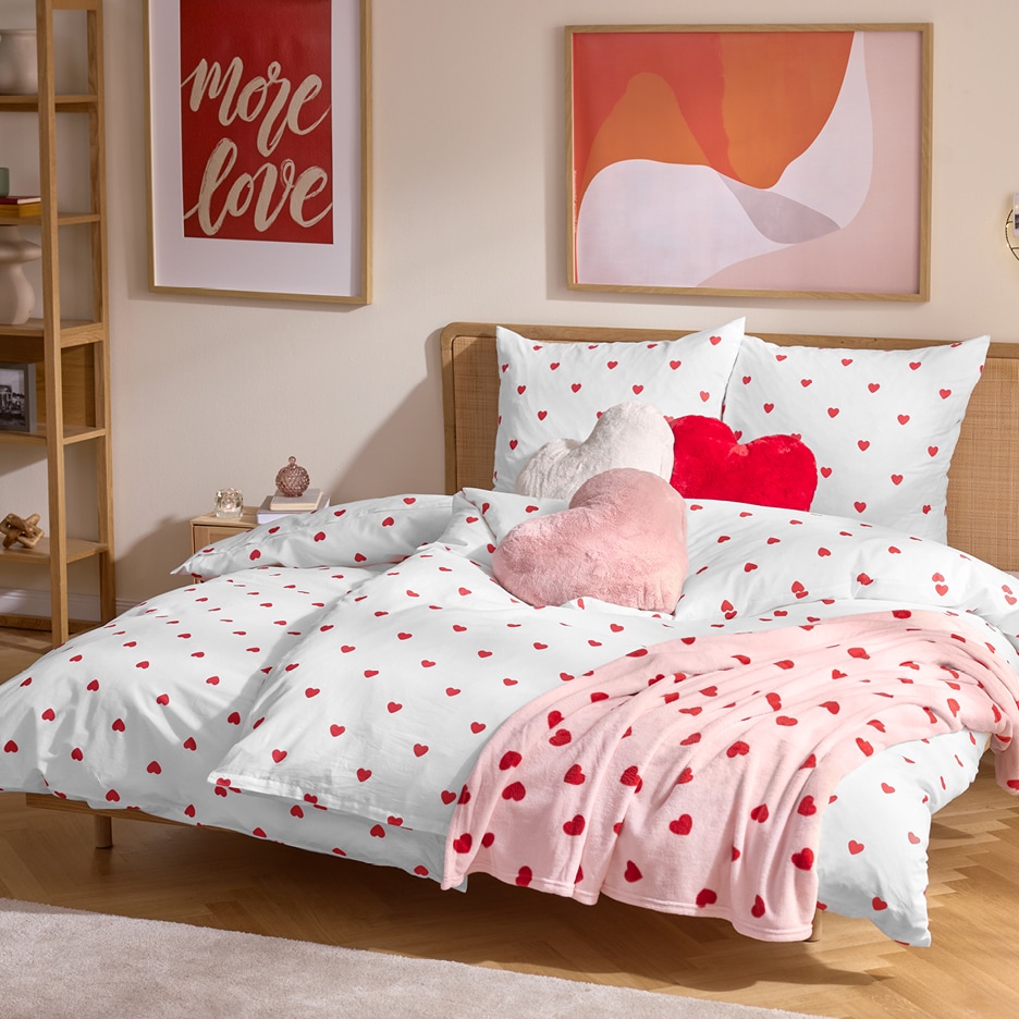 Ložnice s bílým povlečením s červenými srdíčky, růžovou dekou a polštáři ve tvaru srdce.