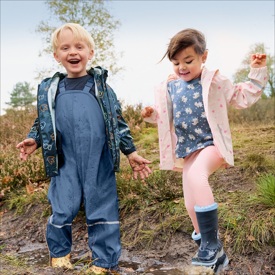 Dvě děti v nepromokavém oblečení a holínkách si hrají v blátě.