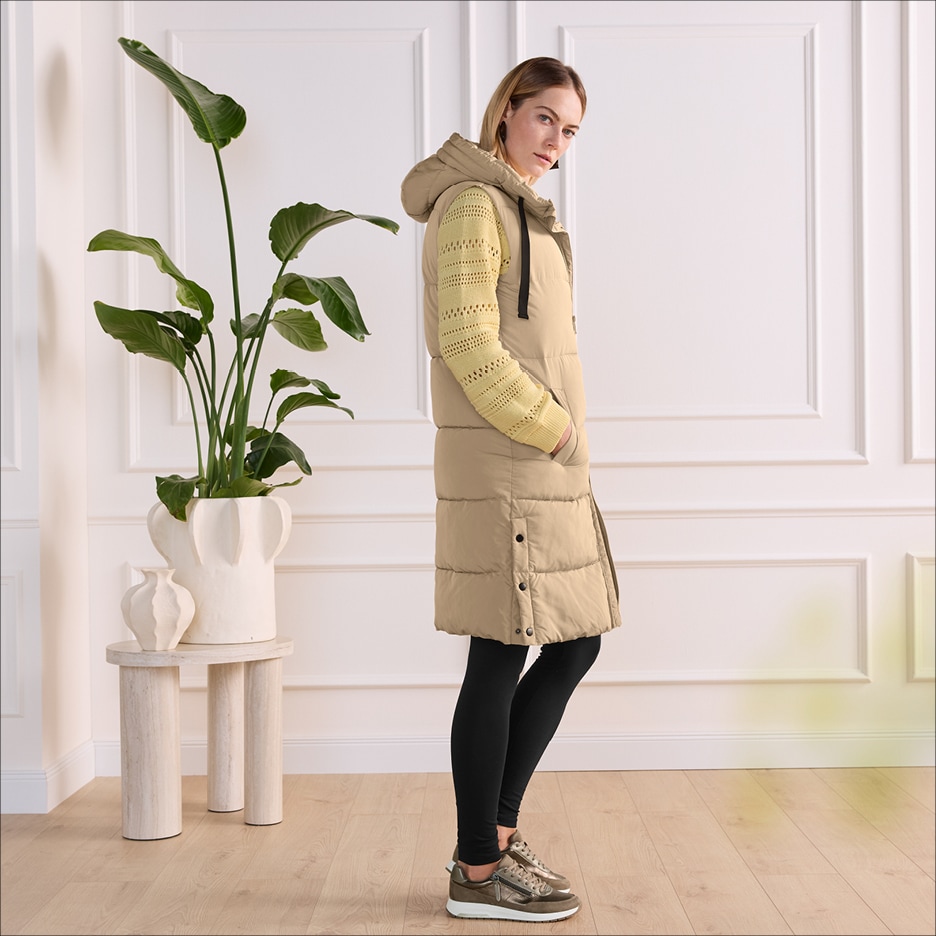 Žena v béžové prošívané vestě, žlutém svetru a černých legínách.