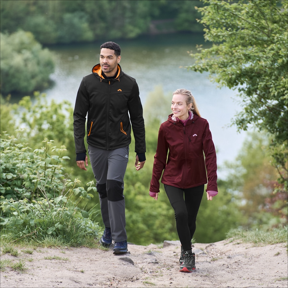 Pár v outdoorovém oblečení, muž v černé bundě a šedých kalhotách, žena v bordó bundě a černých legínách, kráčí po stezce.