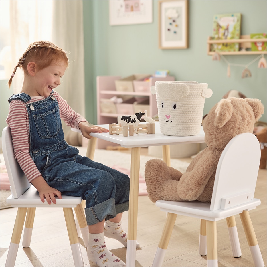 Dítě sedí u bílého dětského stolu s medvídkem a hračkami, v pozadí růžová police.
