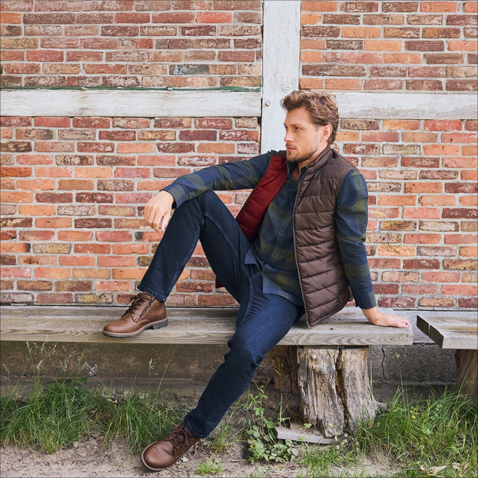 Muž v prošívané vestě, kostkované košili a džínách sedí na dřevěné lavici.