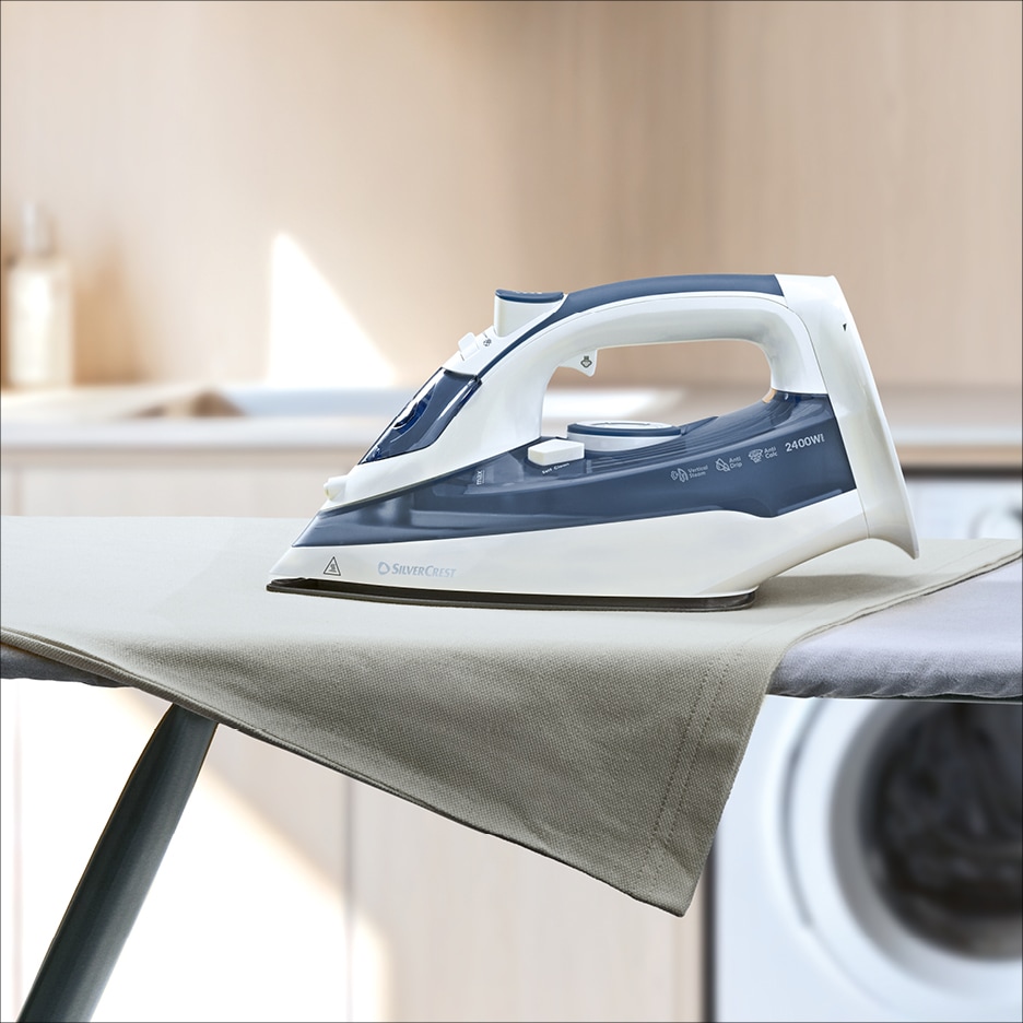 Napařovací žehlička na žehlicím prkně s látkou, v pozadí kuchyně.