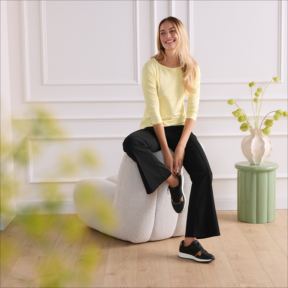 Žena v žlutém tričku s dlouhým rukávem a černých kalhotách sedí na bílém pufu.