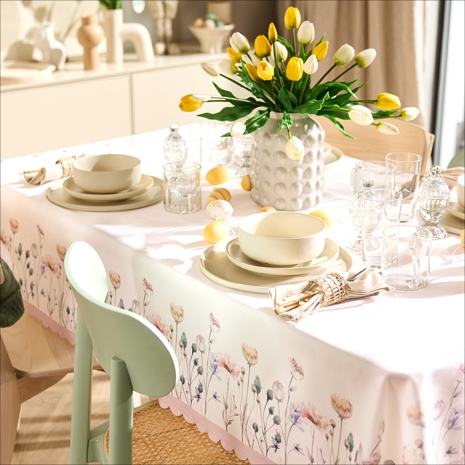Prostřený stůl s jarní výzdobou, tulipány ve váze, nádobí a ubrusem s květinovým vzorem.