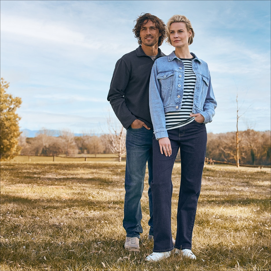 Pár v džínách a ležérním oblečení stojí v trávě s krajinou v pozadí.