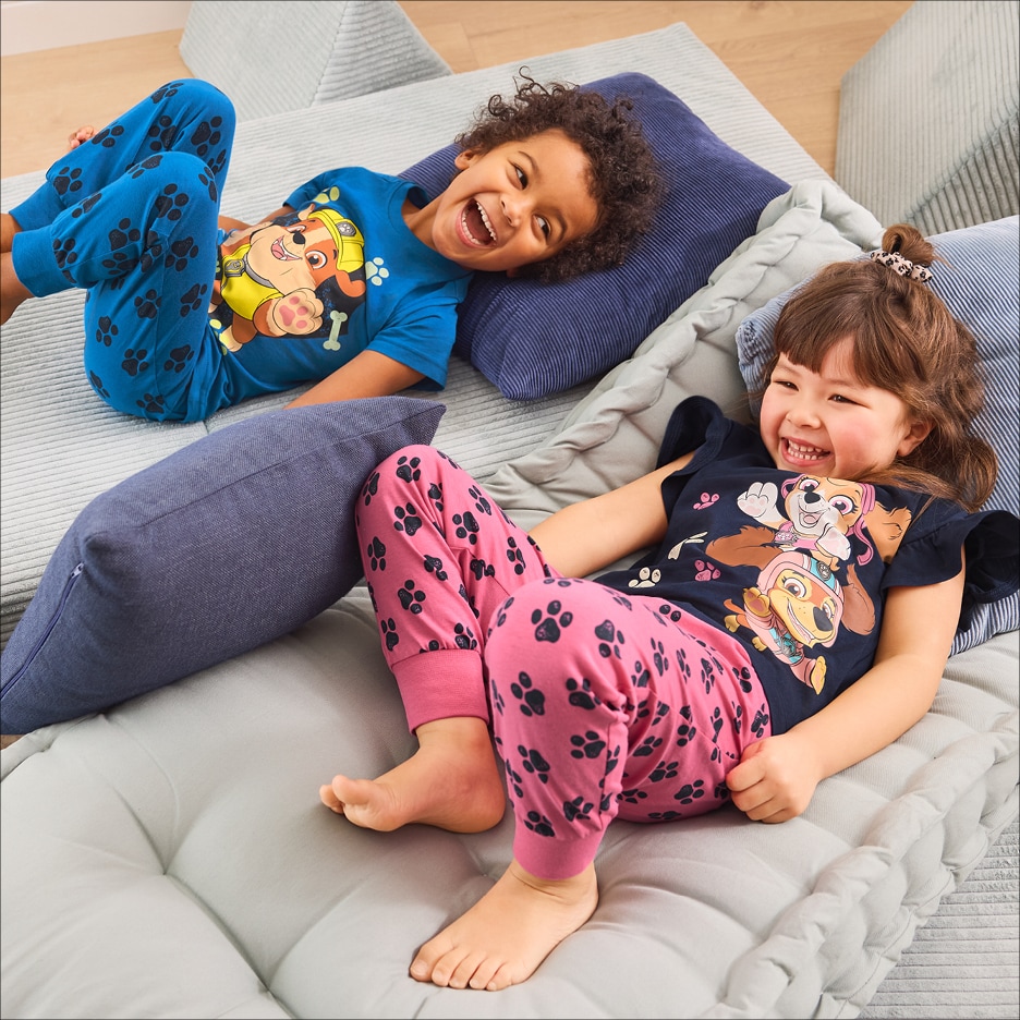 Dvě děti v pyžamech s potiskem tlapek a kreslených psů, ležící na polštářích.