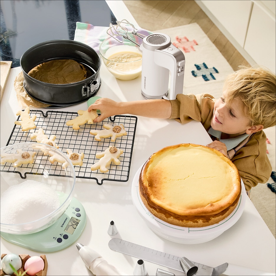 Chlapec zdobí perníčky, na stole je cheesecake, kuchyňská váha a ruční mixér.