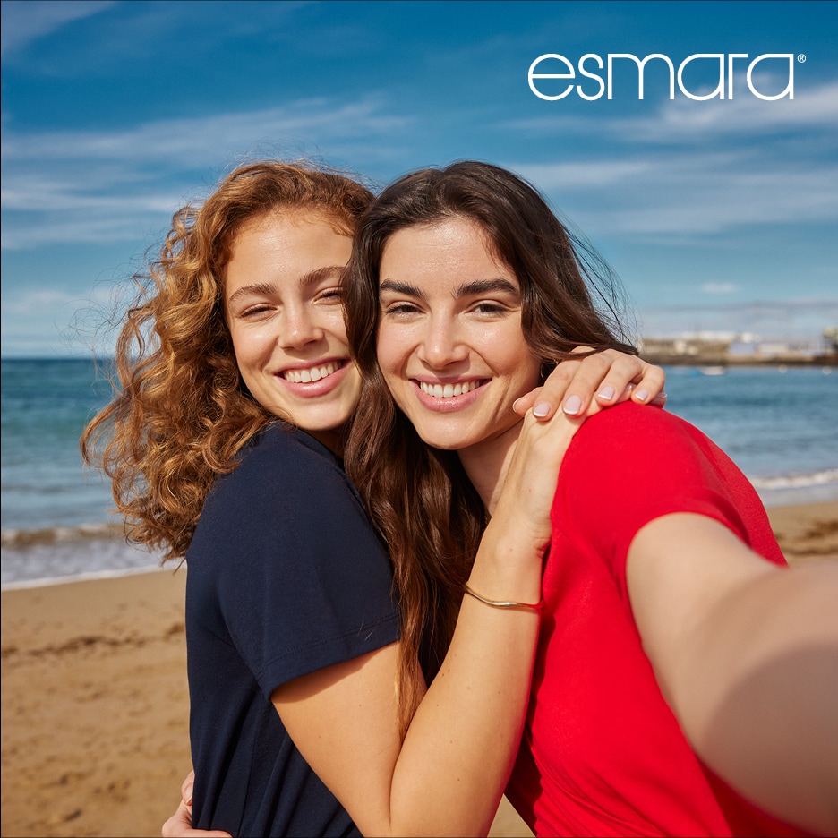 Dvě usměvavé ženy v tričkách se objímají na pláži.