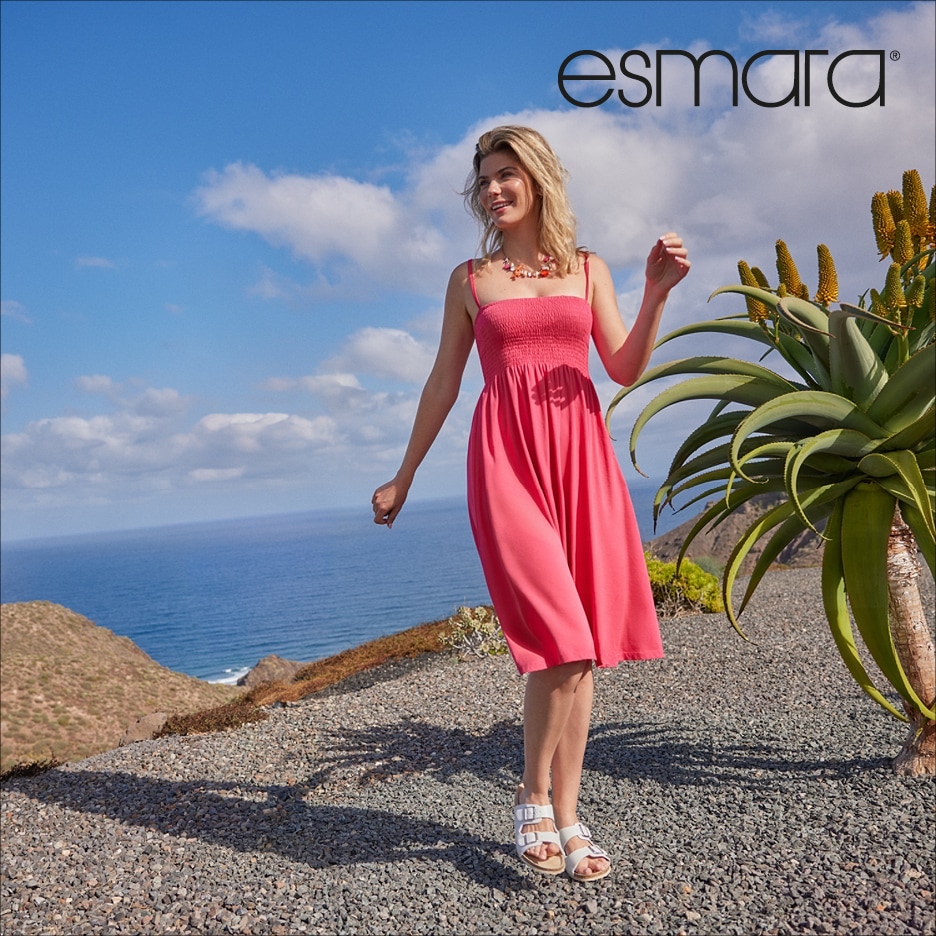 Žena v růžových letních šatech a bílých sandálech stojí na štěrkové cestě s výhledem na moře.