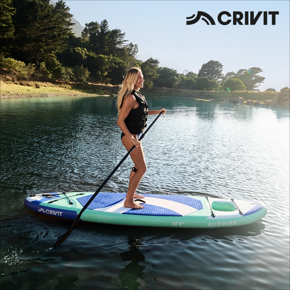 Žena na paddleboardu s pádlem na jezeře, obklopena stromy a horami.