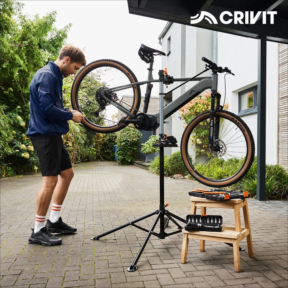 Muž opravuje horské kolo na montážním stojanu, vedle jsou nástroje na dřevěné stoličce.