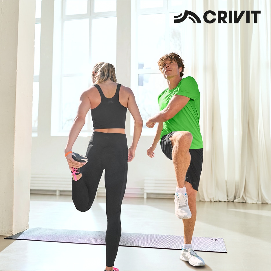 Muž a žena cvičící v aktivním oblečení, žena v černých legínách a sportovní podprsence, muž v zeleném tričku a černých šortkách.