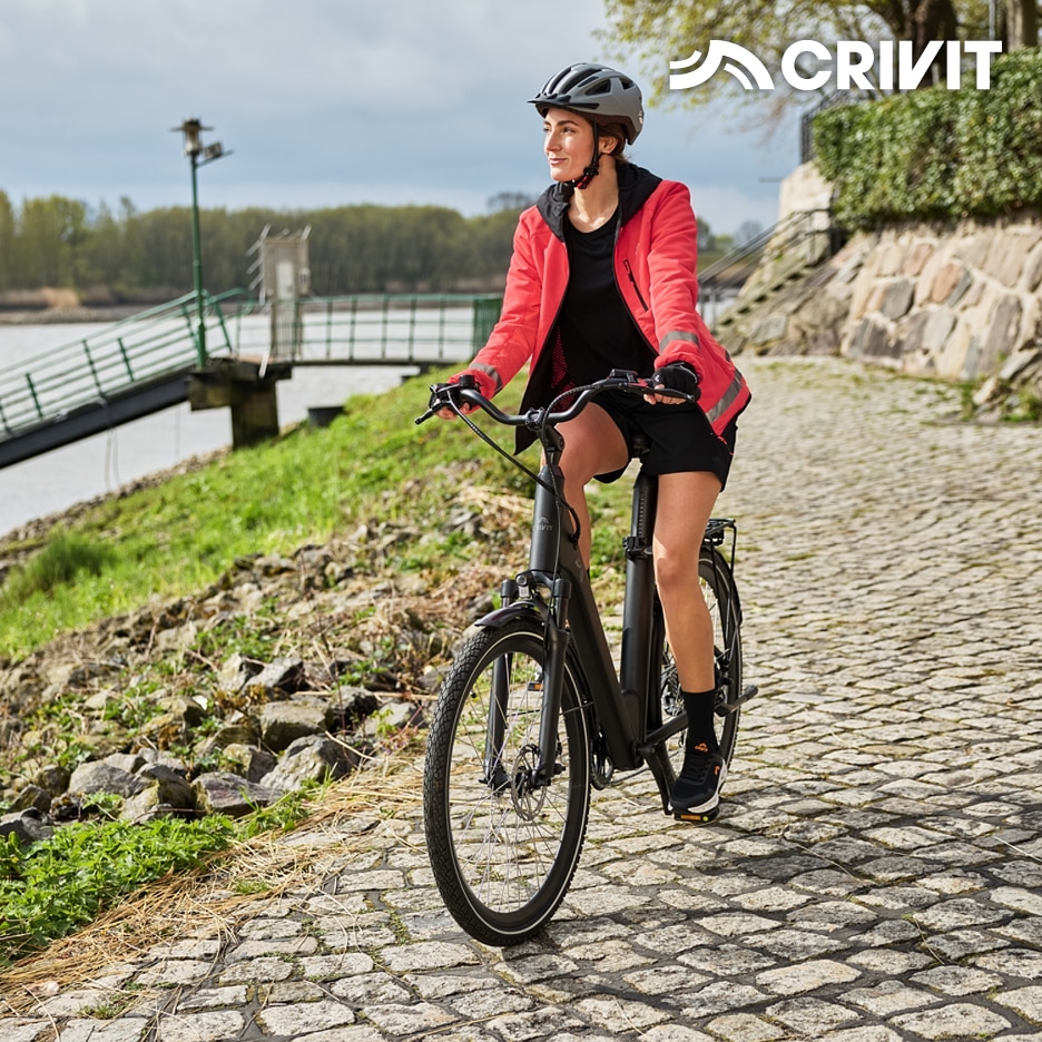 Žena na elektrokole v cyklistickém oblečení a helmě na dlážděné cestě u vody.