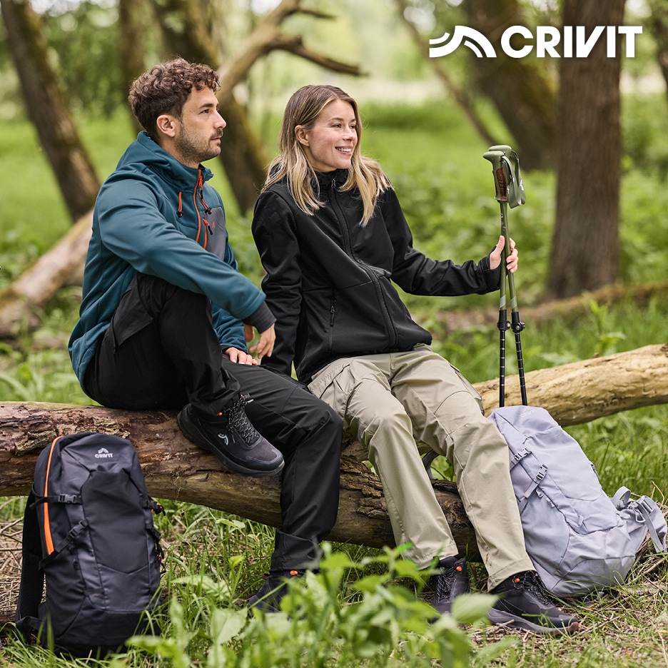 Pár v outdoorovém oblečení sedí na kmeni stromu s batohy a trekovými holemi.