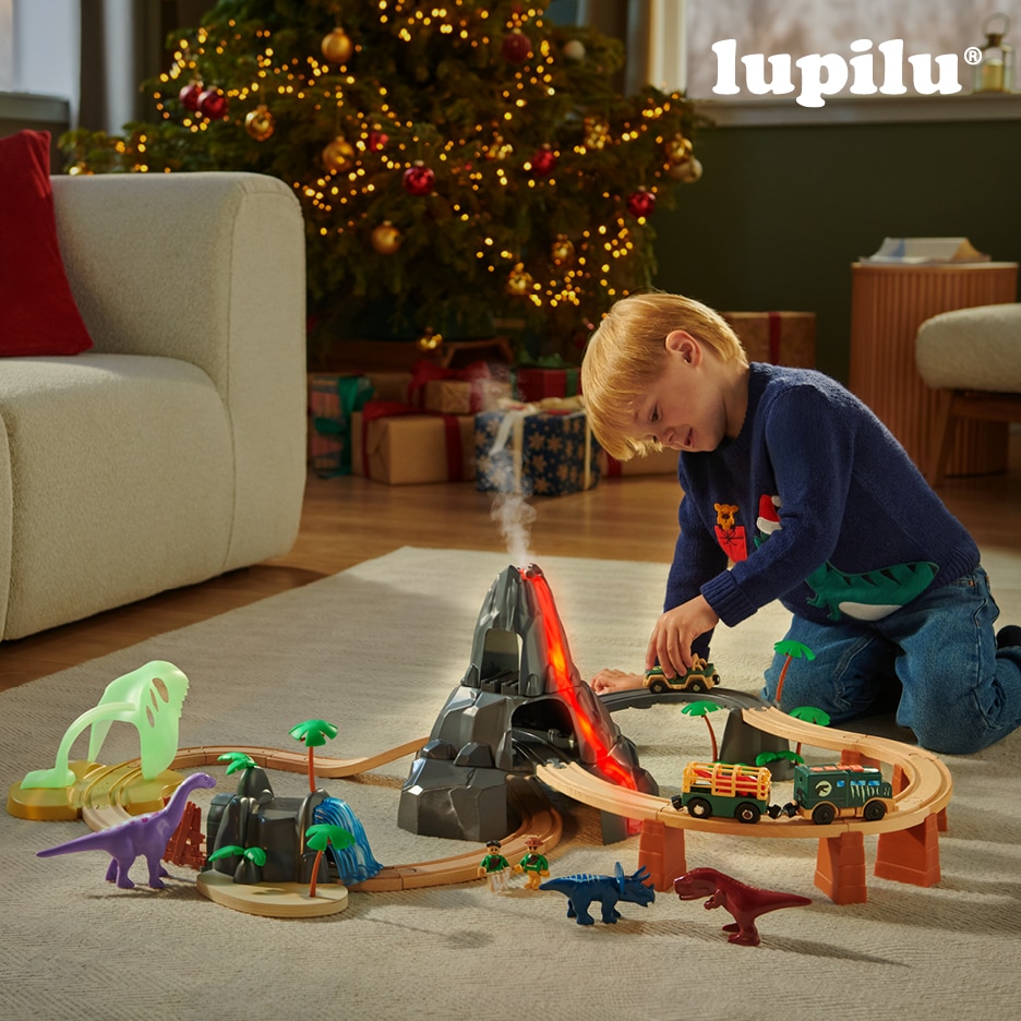 Malý chlapec si hraje s dřevěnou vláčkodráhou s motivem dinosaurů a sopky u vánočního stromku.
