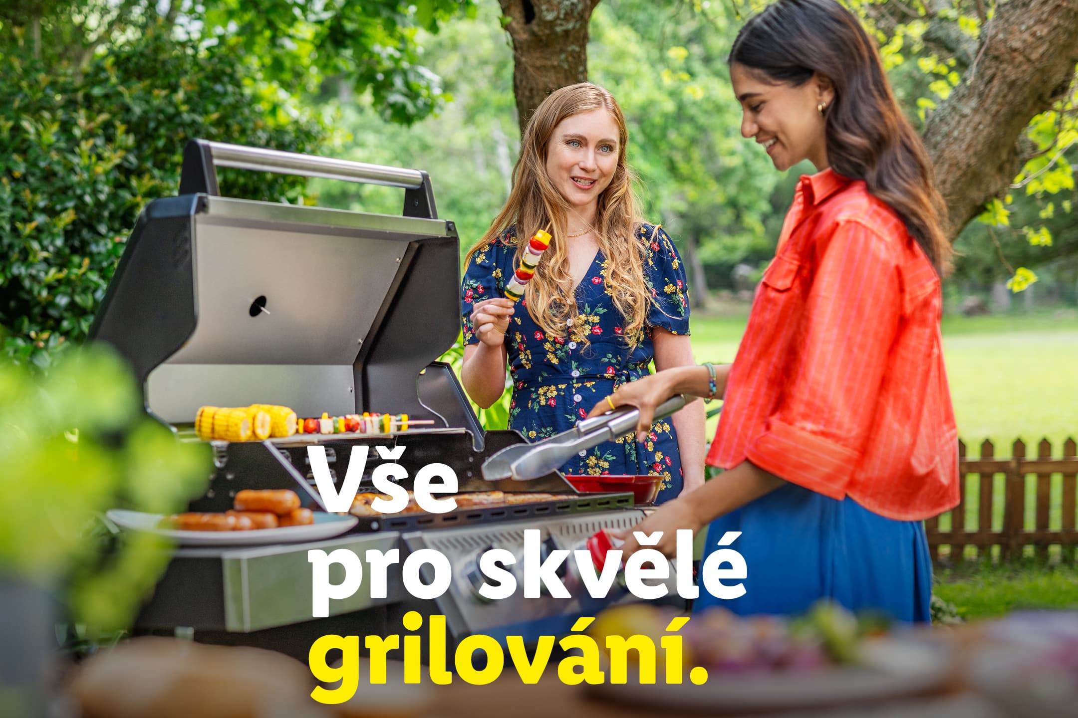 Dvě ženy grilují venku, na grilu jsou klobásy, kukuřice a špízy. Text: Vše pro skvělé grilování.