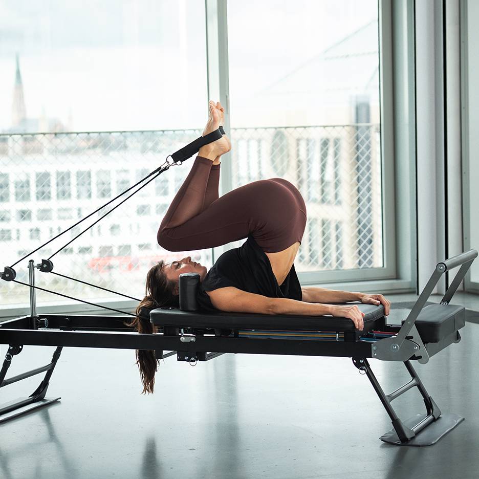 Žena cvičí Pilates na reformer stroji s nohama nahoře a chodidly v popruzích.