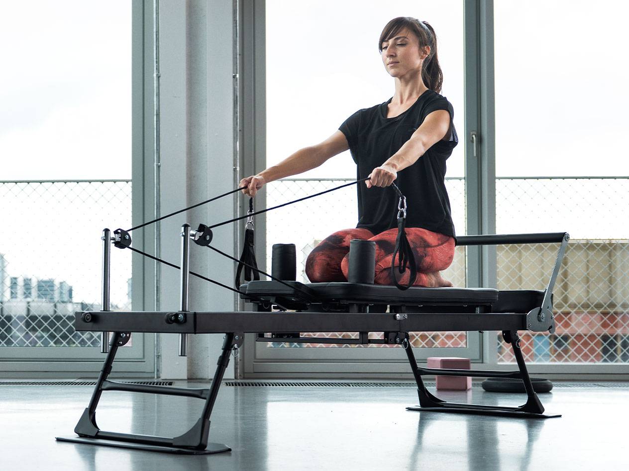 Žena cvičící Pilates na reformeru s velkými okny v pozadí.
