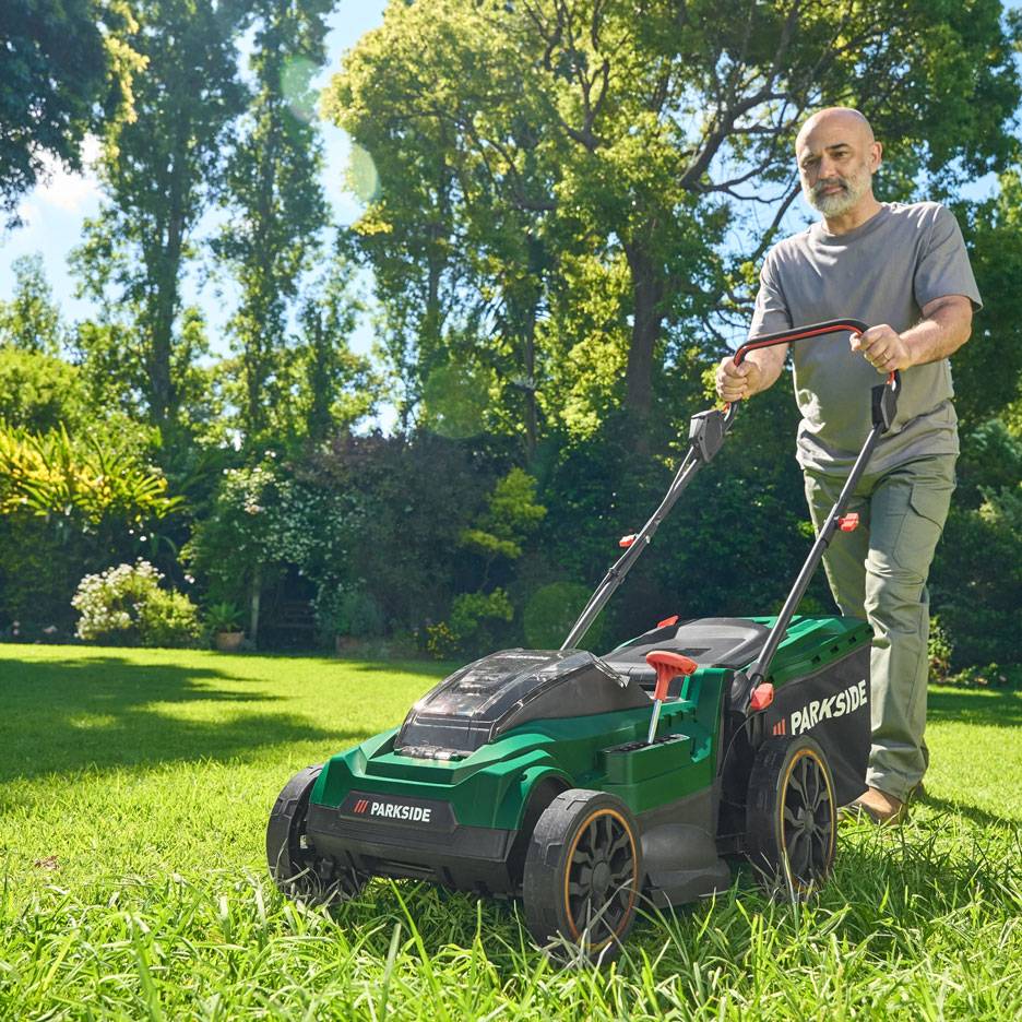 Muž seká trávu zelenou sekačkou v slunné zahradě.